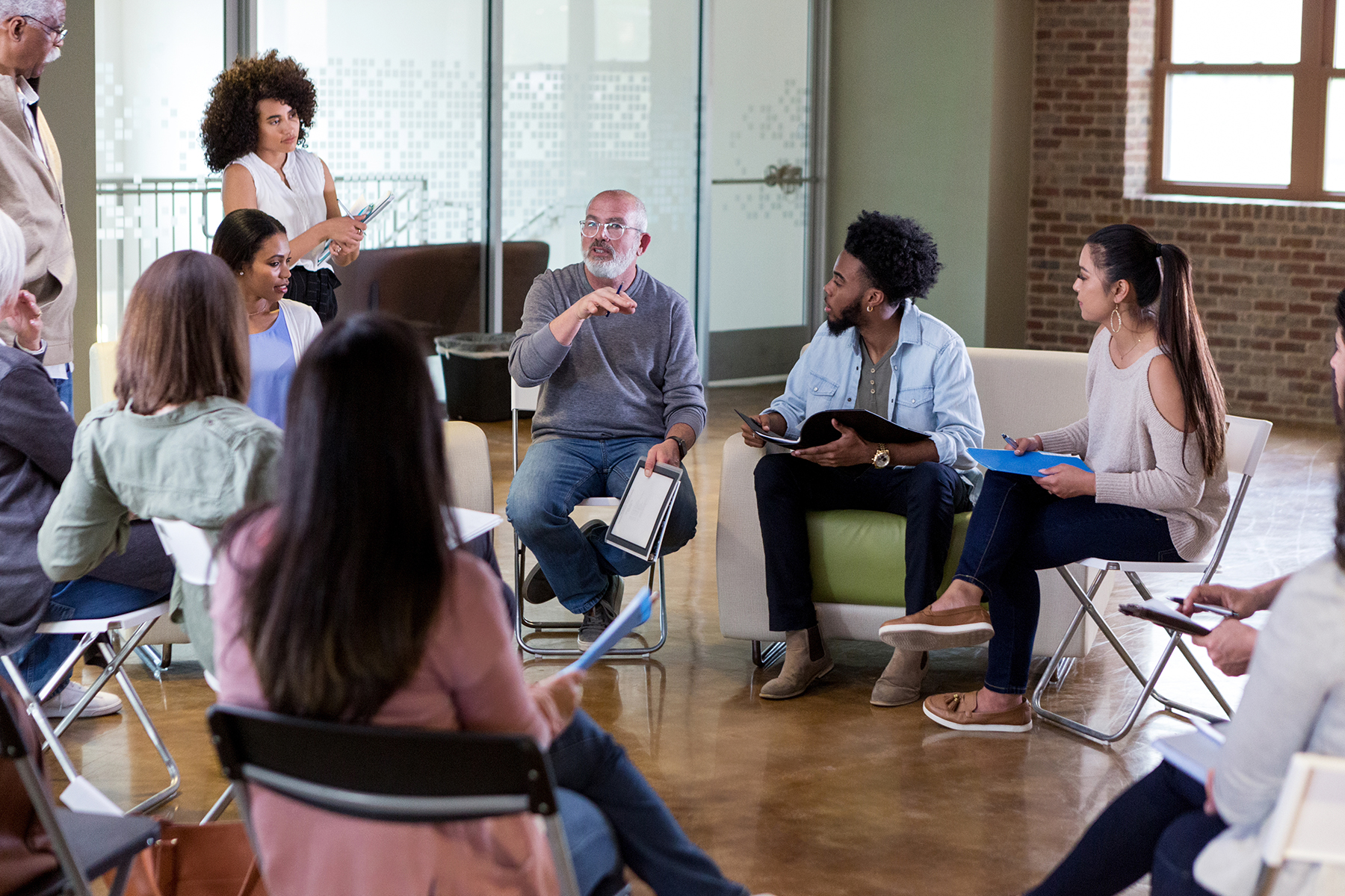 A group of people seated in a circle have a discussion and hold notebooks while a facilitator stands and listens