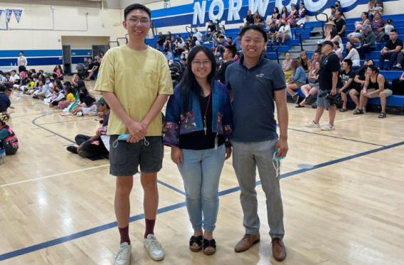 Three smiling people stand facing the camera. They are in a gymnasium and surrounded by bleachers and members of the local Hmong community.